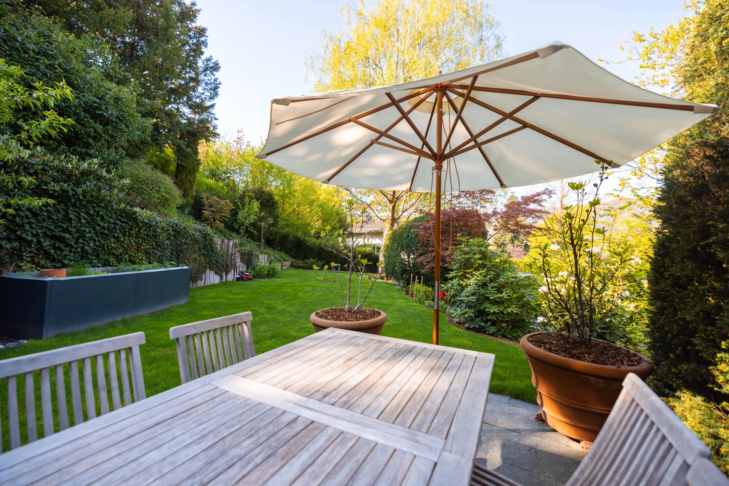 Gepflegter Privatgarten mit Sonnenschirm, Holztisch und Rasenflaeche im warmen Abendlicht, gestaltet von GREGORY Gartenbau